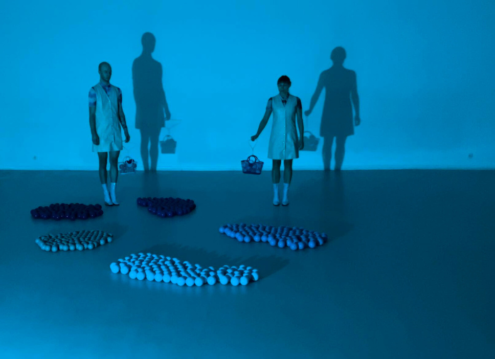 two people / two dancers standing in front of a blue wall in a blue space holding a handbag. there is a sea of 'caviour' on the floor. its a snapshot from a choreography FLARCTAL by Karolina Wyrwal