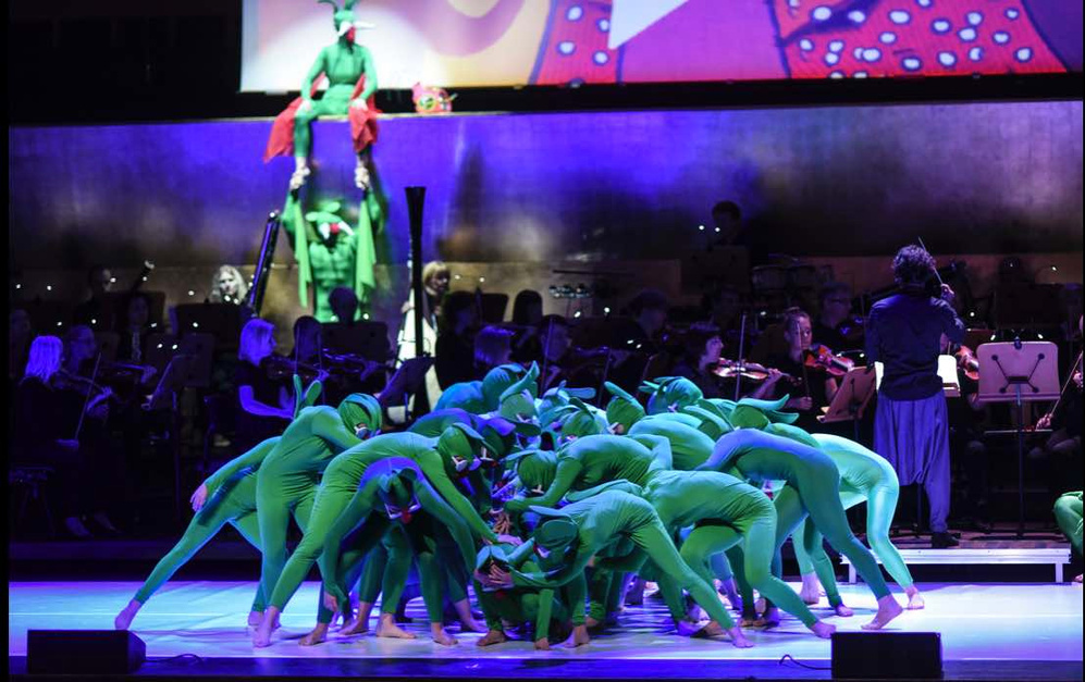 a group of dancers in green costumes performing on stage. there is an orchestra in the background. the picture is from a show Bromba i Muzyka (Szczecin's Phillcharmonic)